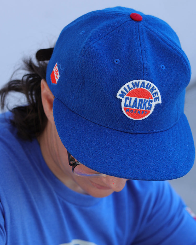 Person wearing the EFF Milwaukee Clarks Vintage Ballcap—a bright blue cap with a red and white logo patch on the front and a small red side tag—styled like a classic IHL wool cap, paired with a matching blue shirt.