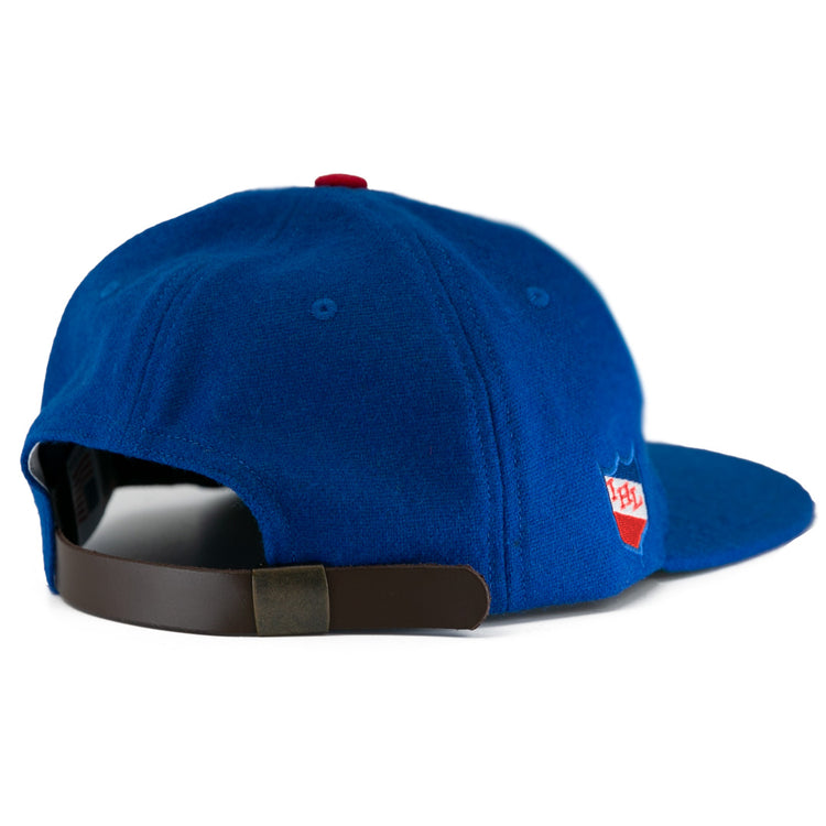 The EFF Milwaukee Clarks Vintage Ballcap is a blue wool cap with a brown adjustable strap and a red-and-white patch on the side, styled like a classic goalkeeper hat. Shown from the rear on a white background.