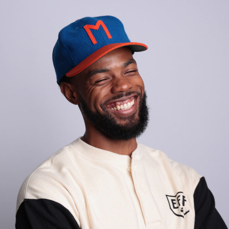 A smiling bearded man wears a blue and orange Morgan State University 1975 Vintage Ballcap by EFF and a cream and black shirt with the EFF logo, standing against a plain light background.