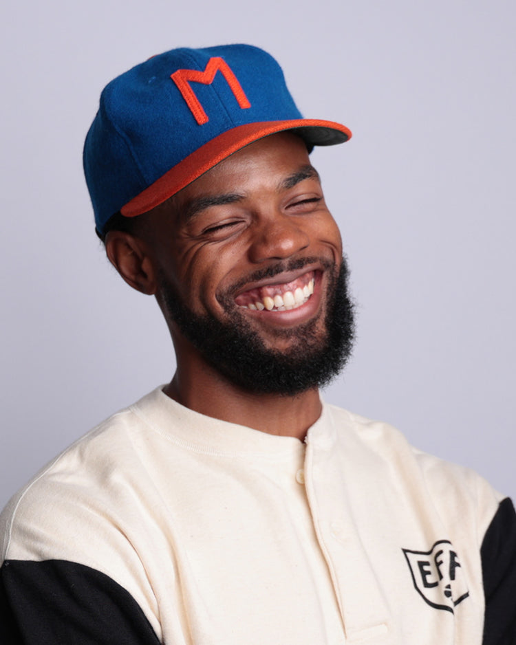 A smiling bearded man wears a blue and orange Morgan State University 1975 Vintage Ballcap by EFF and a cream and black shirt with the EFF logo, standing against a plain light background.
