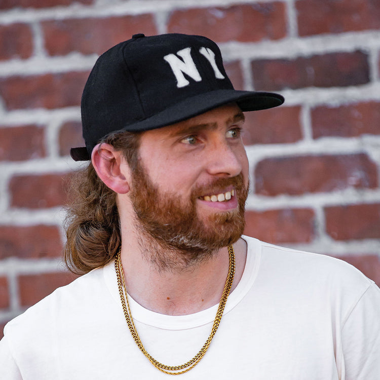 A bearded man smiles in front of a brick wall, wearing an EFF New York Black Yankees Vintage Inspired Ballcap, a white t-shirt, and gold chains.