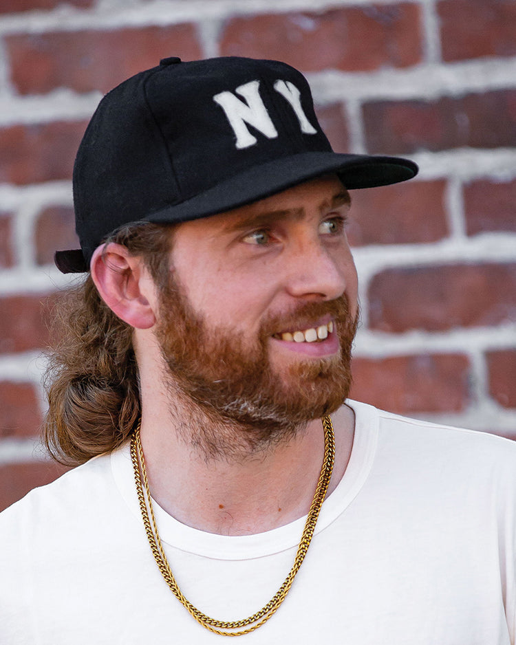 A bearded man smiles in front of a brick wall, wearing an EFF New York Black Yankees Vintage Inspired Ballcap, a white t-shirt, and gold chains.