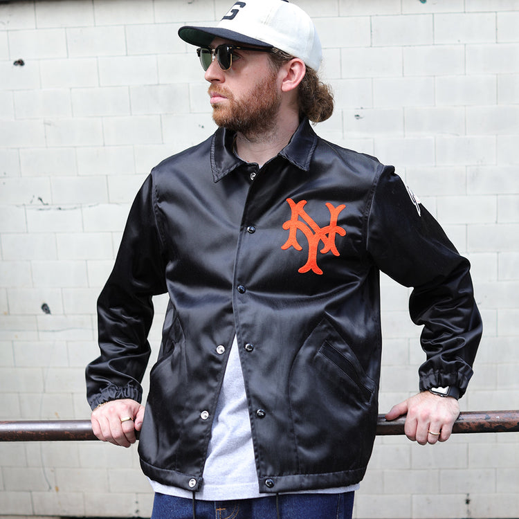 A bearded man in sunglasses, wearing an EFF New York Giants 1954 Satin Windbreaker, a gray cap, and blue jeans, leans casually on a rusted metal railing in front of a tiled wall.