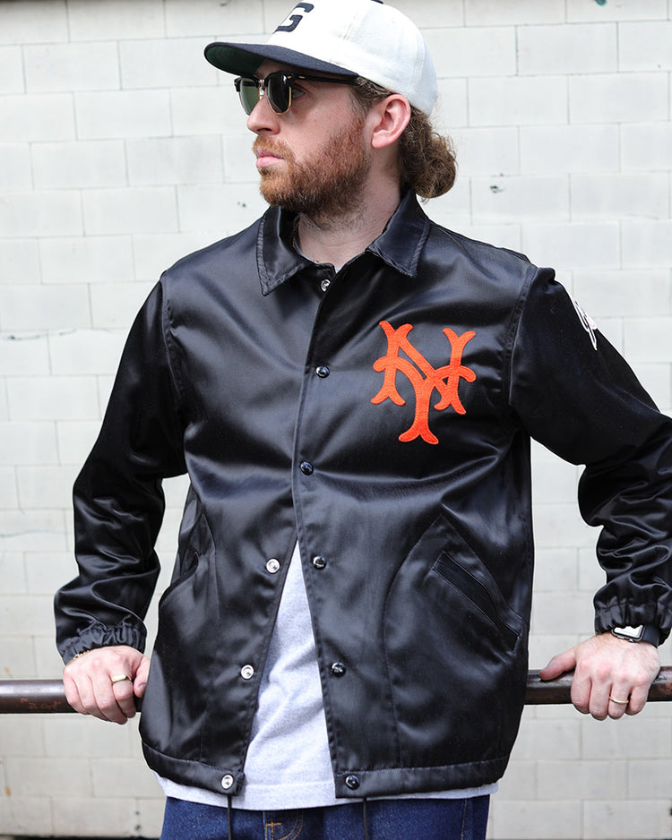 A bearded man in sunglasses, wearing an EFF New York Giants 1954 Satin Windbreaker, a gray cap, and blue jeans, leans casually on a rusted metal railing in front of a tiled wall.