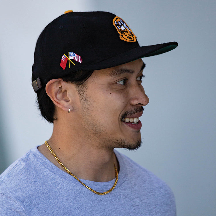 A man in an EFF Pittsburgh Hornets 1953 Vintage Ballcap, gray t-shirt, and gold chain smiles while looking to the side against a neutral background.