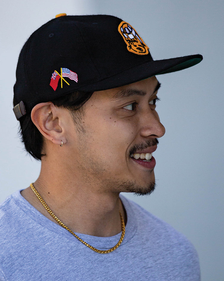 A man in an EFF Pittsburgh Hornets 1953 Vintage Ballcap, gray t-shirt, and gold chain smiles while looking to the side against a neutral background.