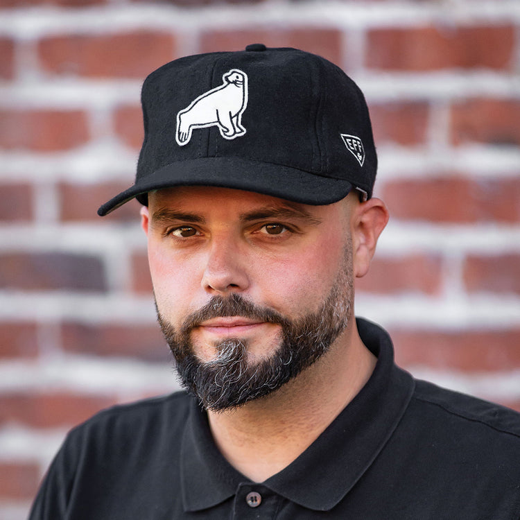A man with a trimmed beard and mustache wears an EFF San Francisco Seals Vintage Inspired Ballcap and a black polo shirt, standing in front of a red brick wall.