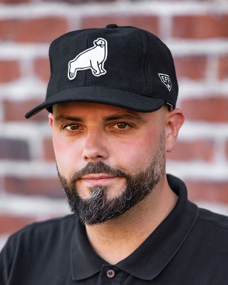 A man with a trimmed beard and mustache wears an EFF San Francisco Seals Vintage Inspired Ballcap and a black polo shirt, standing in front of a red brick wall.