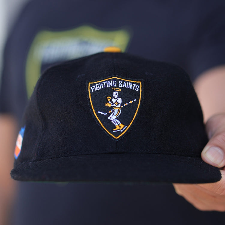 A person holds the EFF St. Paul Fighting Saints 1961 Vintage Ballcap, featuring a cartoon hockey player inside a yellow-bordered shield logo on the front, with a blurred background highlighting the cap.